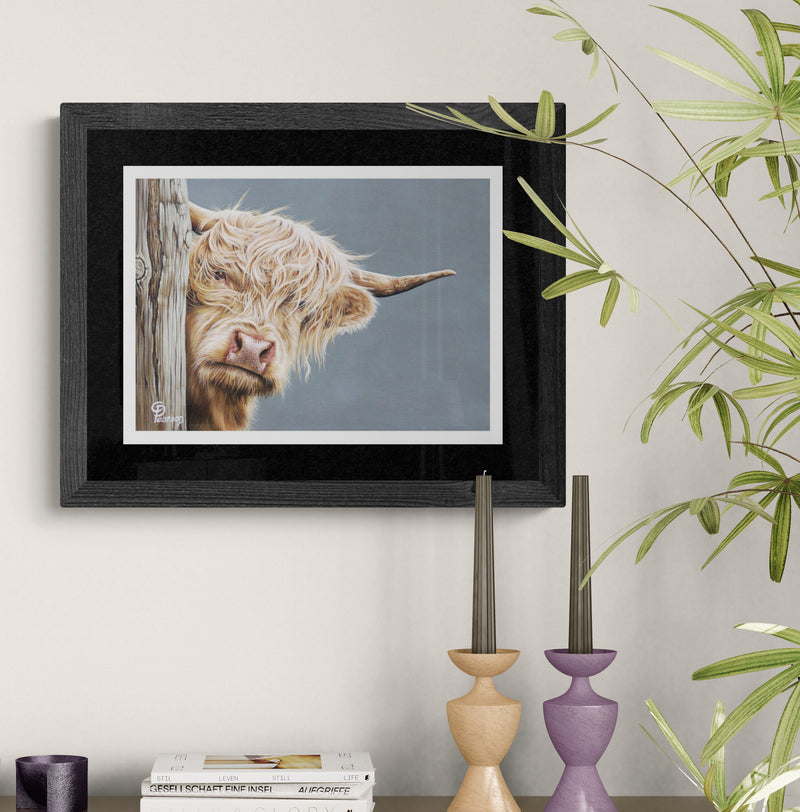 Framed artwork of a Highland cow peeking from behind a log on a wall with decorative elements.