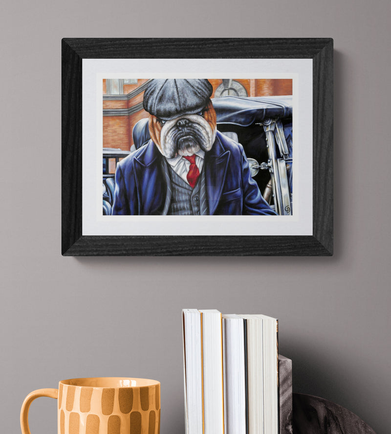 Framed artwork of a bulldog in a suit on a gray wall with books and a mug below.
