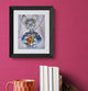 Framed artwork of a cat holding a Rubik's Cube on a pink wall with books and a mug below.