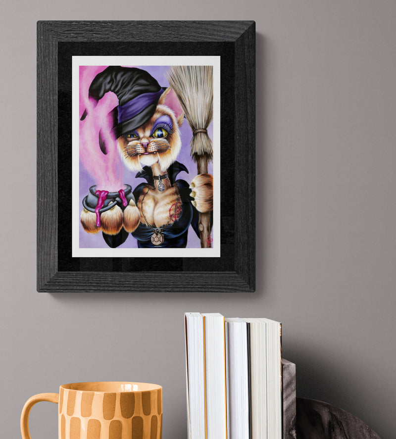 Framed artwork of a cat in a witch costume holding a broom on a gray wall.
