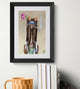 Framed artwork of a cat with a flower and cigarette on a wall above books and a mug.