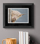 Framed artwork of a highland cow peeking around a wooden post on a gray wall with books and a mug below.
