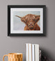 Framed painting of a highland cow on a gray wall with books and a mug below.