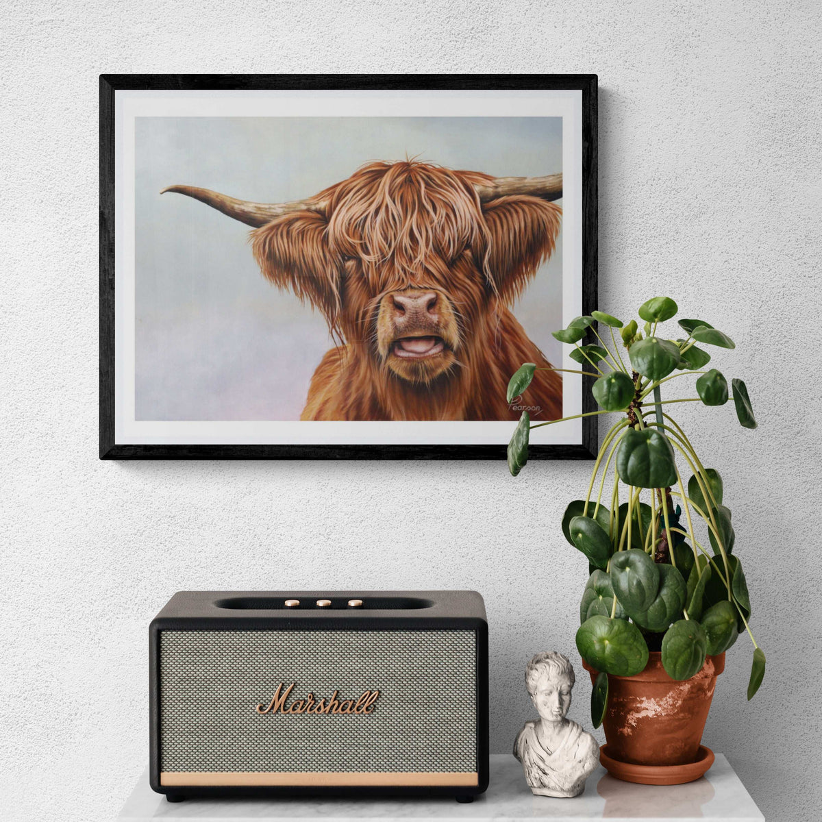 Framed artwork of a Highland cow, Marshall speaker, plant, and small statue on a white surface.