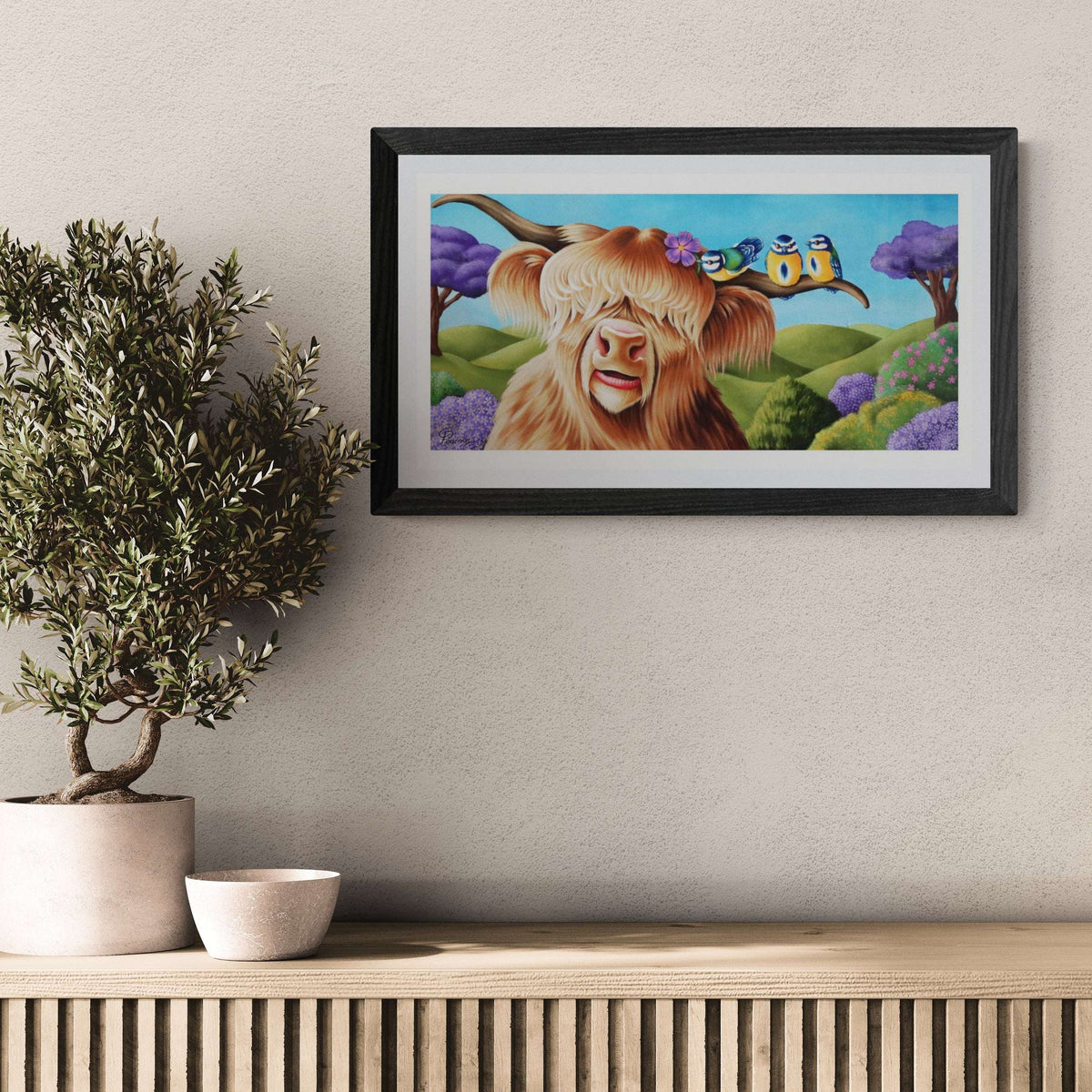Framed artwork of a cow with a butterfly on a wall above a wooden shelf with a plant and bowl.