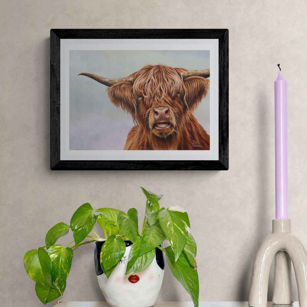 Framed artwork of a highland cow on a wall with a plant and candle in the foreground.