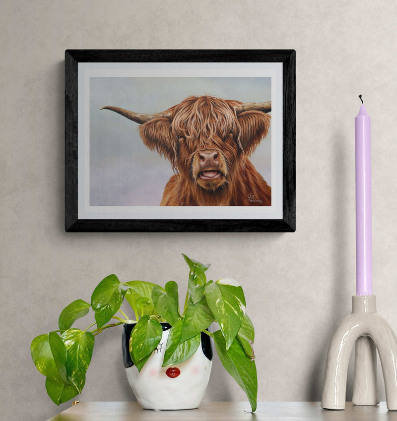 Framed artwork of a highland cow on a wall with a plant and candle in the foreground.