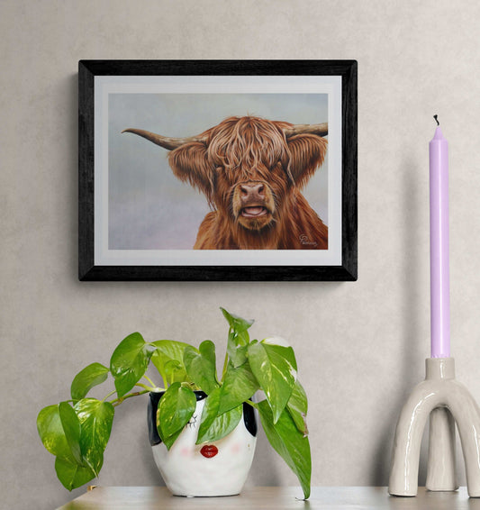 Framed artwork of a highland cow on a wall with a plant and candle in the foreground.