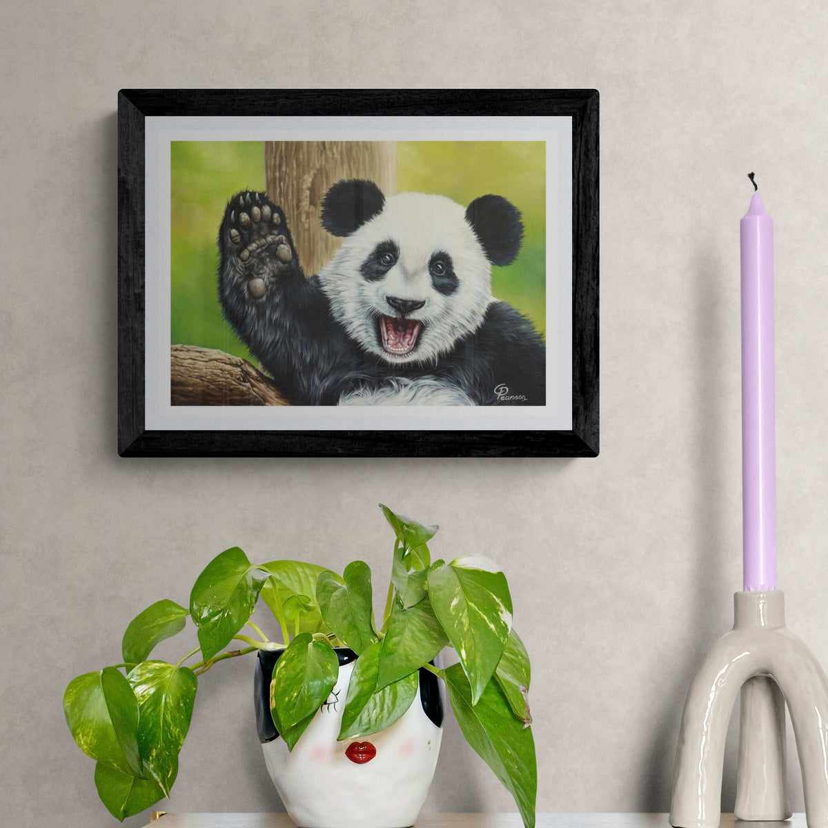 Framed artwork of a panda bear on a wall with a plant and candle in the foreground.