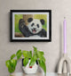 Framed artwork of a panda bear on a wall with a plant and candle in the foreground.