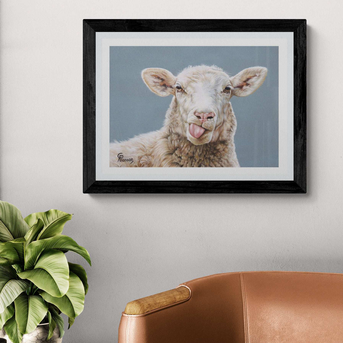 Framed artwork of a sheep with a tongue sticking out on a wall above a brown chair and plant.
