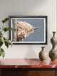 Framed picture of a Highland cow on a wall above a wooden cabinet with vases.