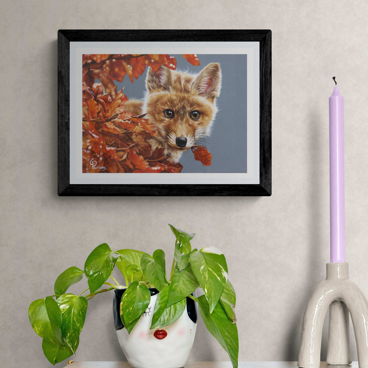 Framed artwork of a fox surrounded by autumn leaves on a wall with decorative items below.
