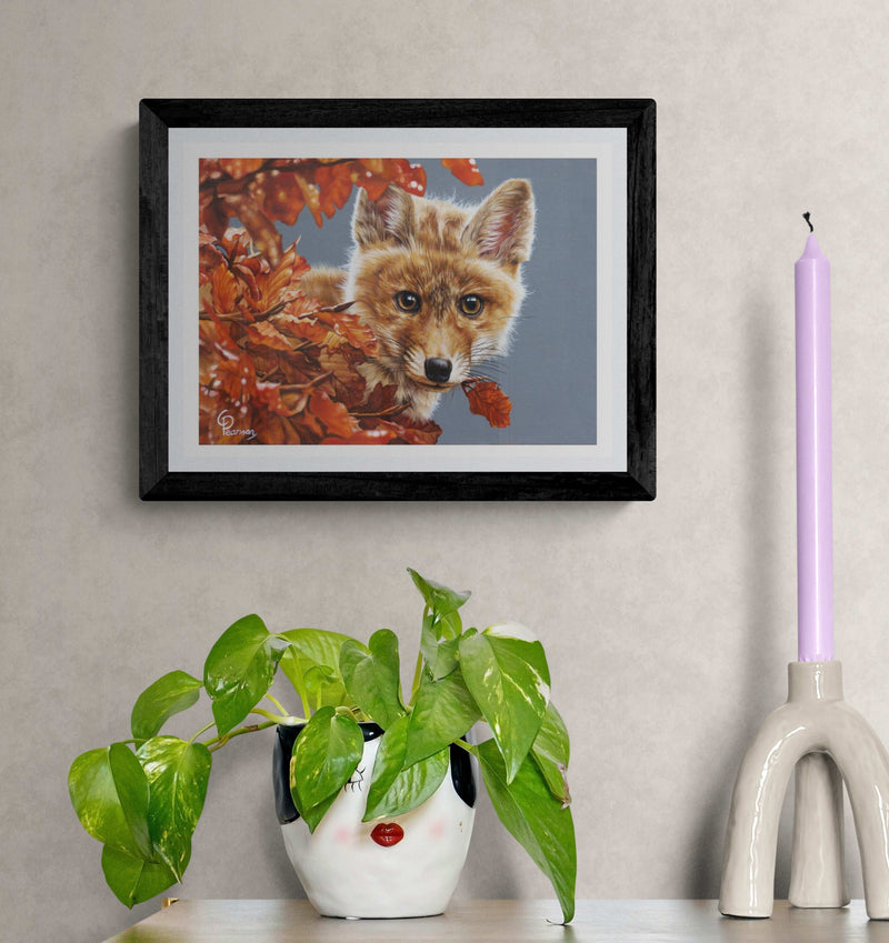 Framed artwork of a fox surrounded by autumn leaves on a wall with decorative items below.