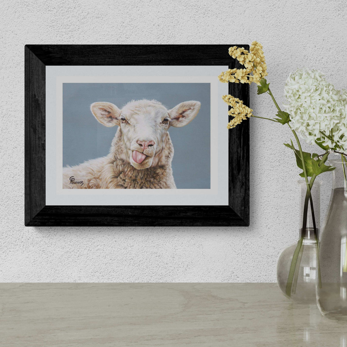 Framed picture of a lamb on a wall with a vase of flowers below.
