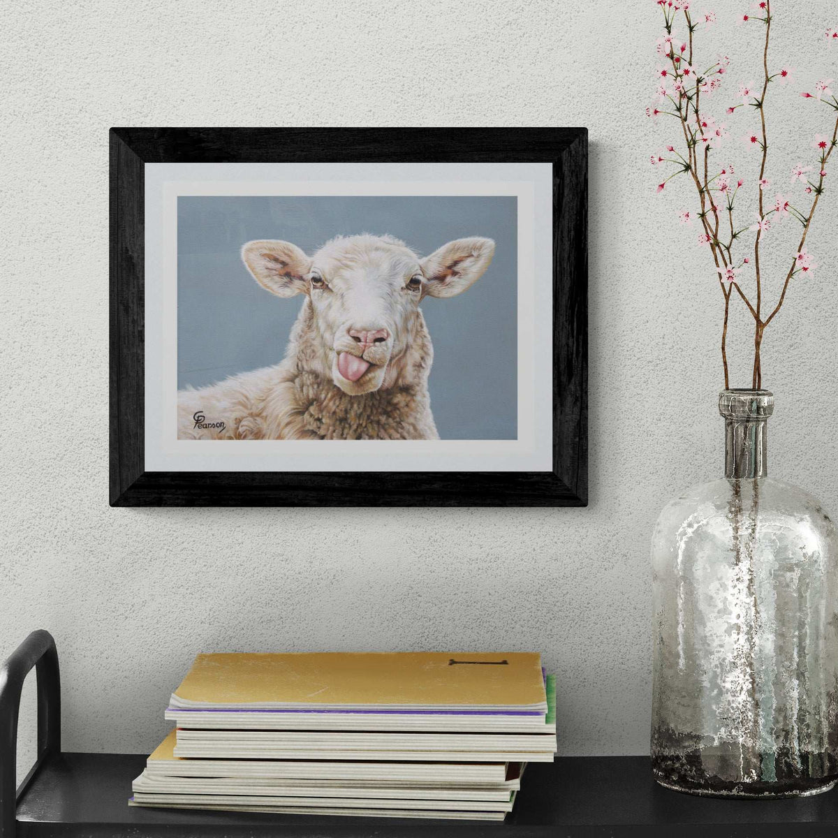 Framed picture of a sheep on a wall above a stack of books with a vase containing branches.