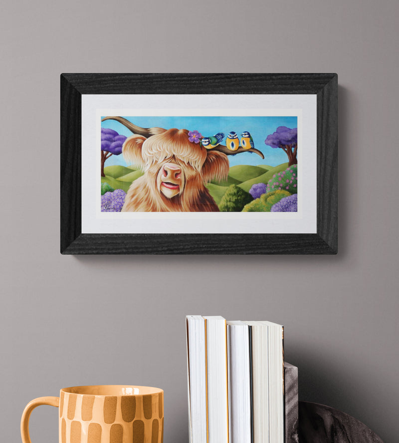 Framed artwork of a cartoonish highland cowwith a colorful landscape on a gray wall.