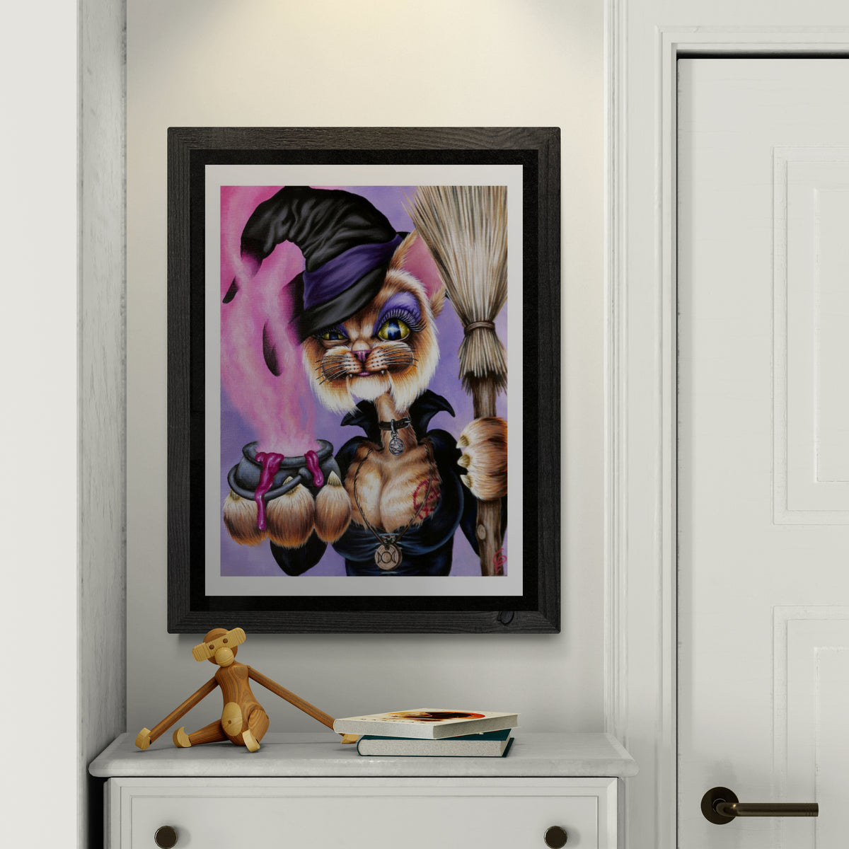 Framed artwork of a cat in a witch costume on a white wall.