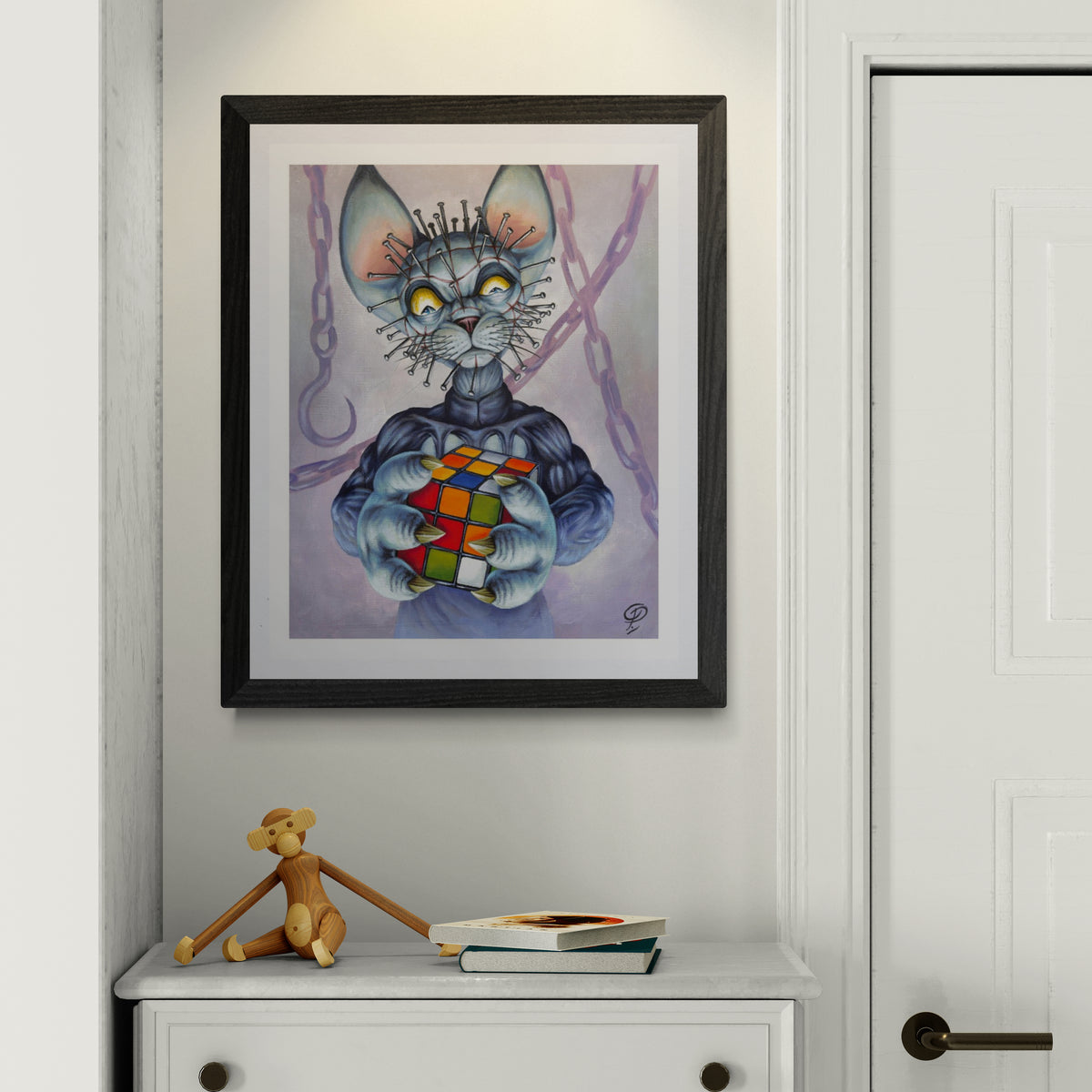 Framed artwork of a cat with a Rubik's Cube on a white wall above a cabinet.