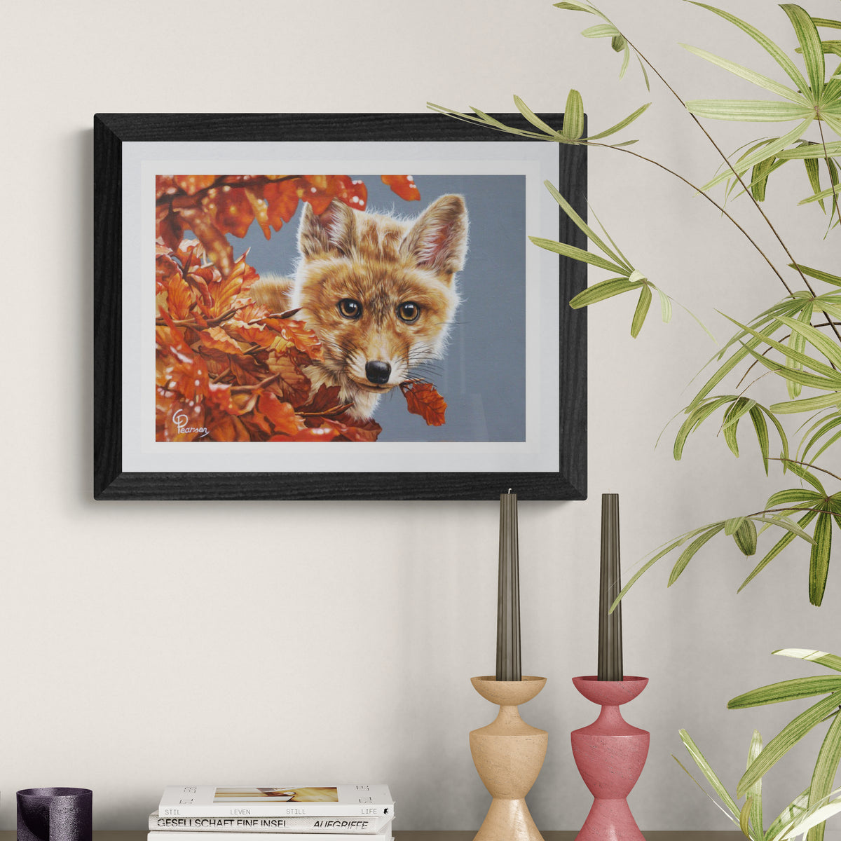 Framed artwork of a fox among autumn leaves on a wall with decorative candles and books.