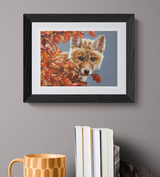 Framed artwork of a fox surrounded by autumn leaves on a gray wall with books and a mug below.