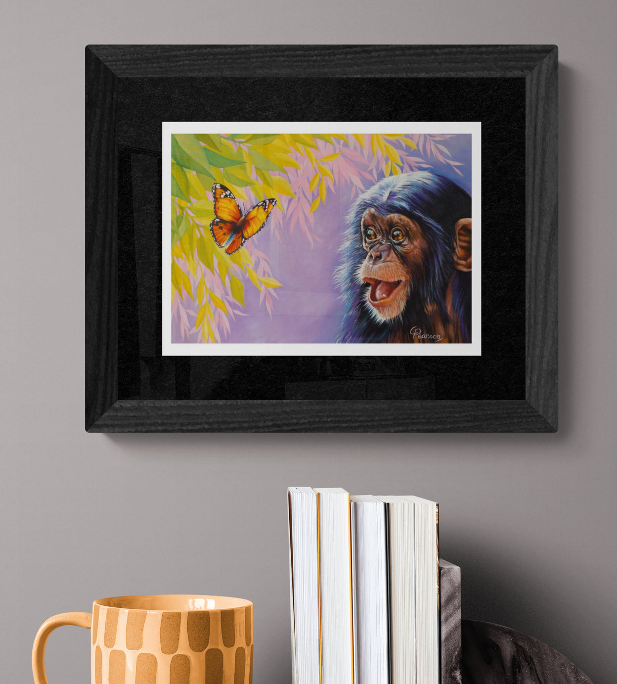 Framed artwork of a monkey and butterfly on a gray wall with books and a mug below.