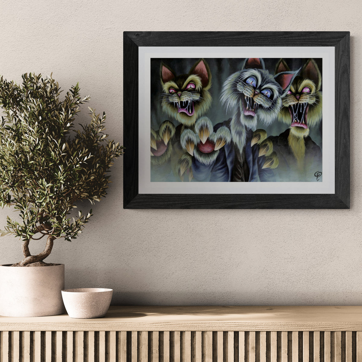 Framed artwork of three cartoon-like zombie cats on a wall above a wooden shelf with a plant and bowl.
